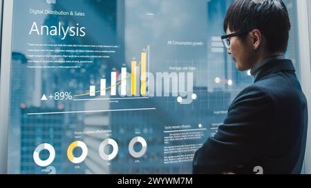 Eleganter japanischer Geschäftsmann, der Unternehmensdiagramm mit dem futuristischen fortschrittlichen holographischen Bildschirm im Aufzug analysiert. Succesful Male fährt zum Büro im modernen Buisness Center. Stockfoto