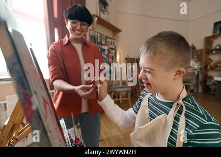 Nahaufnahme Porträt eines glücklichen Jungen mit Behinderung, der an der Staffelei steht und den Pinsel hält, der Kunstunterricht mit einem lächelnden Lehrer genießt Stockfoto