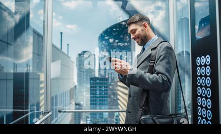 Erfolgreicher Geschäftsmann, der mit dem Glasaufzug zum Büro im modernen Business Center fährt. Gutaussehender Happy man lächelt, während er das Smartphone nutzt, Textnachrichten verfasst, soziale Medien und geschäftliche E-Mails im Handumdrehen durchsucht. Stockfoto