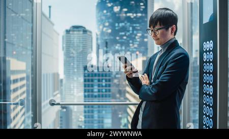 Erfolgreicher, stilvoller japanischer Geschäftsmann, der mit dem Glasaufzug zum Büro im modernen Business Center fährt. Gutaussehender Mann lächelt während der Nutzung des Smartphones, schreibt Textnachrichten, sucht soziale Medien im Handumdrehen. Stockfoto