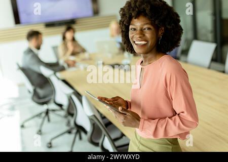 Ccheerful Afroamerikanerin leitet ein Meeting und hält sich an einem digitalen Tablet fest, während Kollegen hinter ihr Gespräche führen. Stockfoto