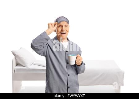 Älterer Mann im Pyjama, der eine Schlafmaske auszieht und eine Tasse isoliert auf weißem Hintergrund hält Stockfoto