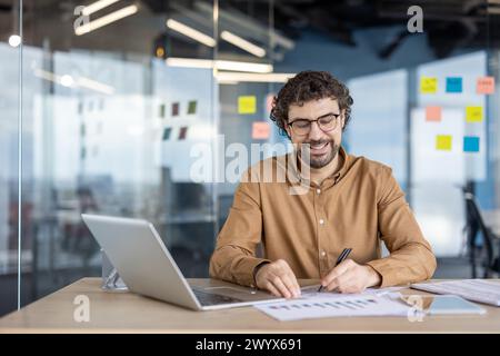 Glücklicher reifer hispanischer Mann, der seine Arbeit mit Laptop und Dokumenten in einer modernen Büroumgebung genießt. Positives Arbeitsumfeld und Konzept der Zufriedenheit am Arbeitsplatz. Stockfoto