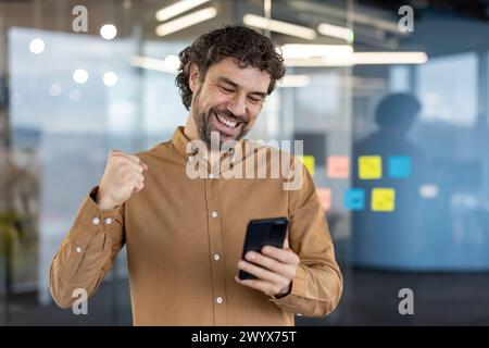 Der fröhliche männliche Unternehmer wirft aufgeregt die Faust in den Sieg, während er sein Smartphone in einem modernen Büroumfeld betrachtet. Stockfoto