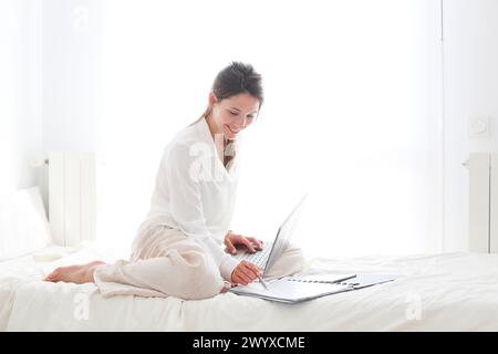 Frau, die auf dem Bett sitzt und einen Laptop benutzt. Stockfoto