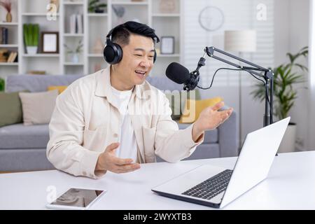 Glücklicher asiatischer männlicher Podcaster, der Audioinhalte zu Hause mit Laptop und Studiomikrofon aufnimmt und damit das Online-Publikum anspricht. Stockfoto