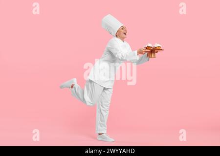 Glücklicher professioneller Konditor in Uniform, der Cupcakes auf rosa Hintergrund hält Stockfoto