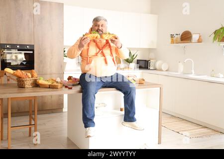 Übergewichtiger reifer Mann, der auf einem Tisch voller ungesunder Lebensmittel in der Küche sitzt. Das Konzept des Überessens Stockfoto