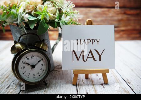 Happy May Textnachricht auf Papierkarte mit Holzstaffelei und Wecker mit Blume in Metallvase Dekoration Stockfoto