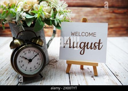 Begrüßungsnachricht im August auf Papierkarte mit Holzstaffelei und Wecker mit Blume in Metallvase Stockfoto