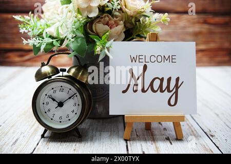 Begrüßungsnachricht im Mai auf Papierkarte mit Holzstaffelei und Wecker mit Blume in Metallvase Stockfoto