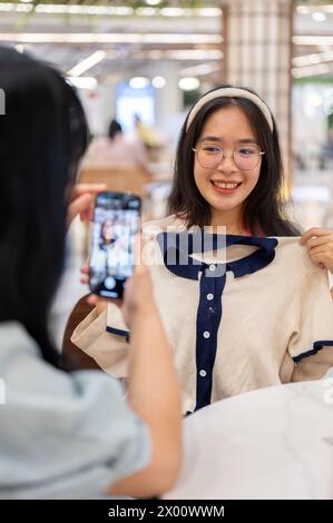 Zwei glückliche junge Frauen sitzen in einem Café in einem Einkaufszentrum und haben gemeinsam einen lustigen Einkaufstag. Eine Frau, die ein Foto von ihrem Freund mit einem neuen Clothe macht Stockfoto