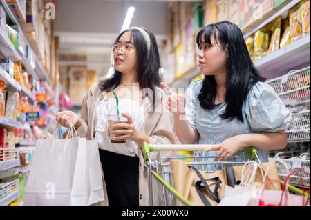 Zwei junge asiatische Frauen kaufen zusammen in einem Supermarkt oder Supermarkt ein, wobei sie sich auf die Auswahl von Artikeln aus den Regalen konzentrieren und mit einem Einkaufswagen laufen. Einkaufen Stockfoto