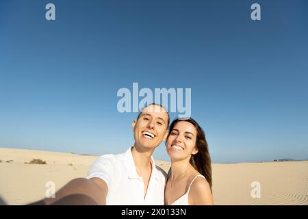 Fröhliches junges Paar, das in die Kamera blickt, während es auf sandigem Gelände bei sonnigem Tageslicht steht und Selfie mit zahnlosem Lächeln gegen wolkenlose blaue s macht Stockfoto