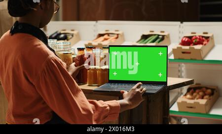 Der junge Anbieter betrachtet den Greenscreen auf dem Notebook, während er die Waren für Kunden vorbereitet, indem er ein isoliertes Modell-Layout auf dem Bildschirm verwendet. Verkäufer in afroamerika überprüft leere Chromakey-Anzeige. Kamera 1. Stockfoto