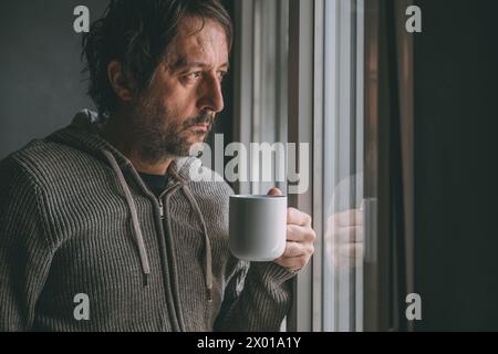 Nachdenklicher männlicher Erwachsener trinkt Kaffee am Morgen am Fenster des Appartements, während er hinausschaut und nachdenkt. Coffeindosis zum Tagesbeginn. Wählen Sie Aus Stockfoto