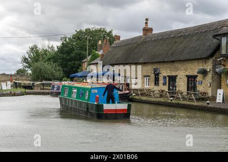 Schmalboot navigiert durch den Grand Union Kanal mit dem Boat Inn im Hintergrund, Stoke Bruerne, Northamptonshire, Großbritannien Stockfoto