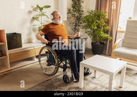 Ein älterer Mann sitzt zu Hause im Wohnzimmer im Rollstuhl und lernt, wie man Schlagzeug spielt, indem er online auf einem Laptop unterrichtet. Stockfoto