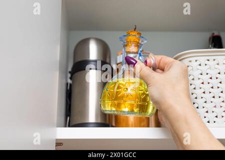 Die Hausfrau nimmt eine Glasflasche Sonnenblumenöl aus dem Küchenschrank. Glasflasche mit Speiseöl. Olivenölflasche. Ein Mann hält eine Flasche davon Stockfoto