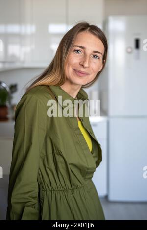 Lächelnde skandinavische Frau mittleren Alters mit zufriedenem Ausdruck, die in der Küche zu Hause in die Kamera schaut Stockfoto