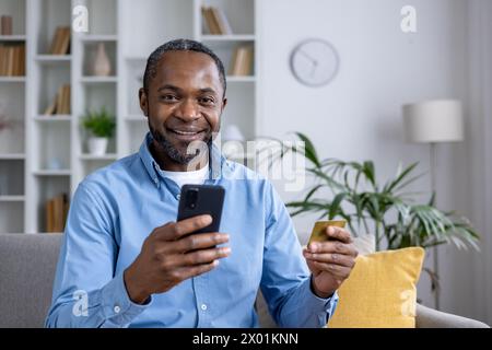 Fröhlicher afroamerikanischer Mann zu Hause, der die Kamera anlächelt, während er ein Smartphone und eine Kreditkarte hält und Online-Shopping oder -Banking porträtiert. Stockfoto