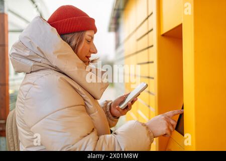 Lächelnde schöne Kaukasierin Klientin mit Pappkartons und Händen zeigen Smartphone mit automatisierter Selbstbedienungs-Post-Terminal-Maschine oder Stockfoto