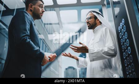 Geschäftsmann spricht mit Arab Investment Partner, während er mit dem Glasaufzug zum Büro in einem modernen Business Center fährt. Internationale Corporate Associates diskutieren Details eines Deals in einem Lift. Stockfoto