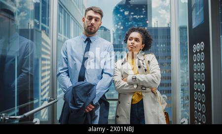 Zwei Bürokollegen, die auf dem Weg von der Arbeit im Glasaufzug in einem modernen Business Center unterwegs sind. Kaukasischer männlicher Spezialist und schwarzer lateinamerikanischer weiblicher Manager plaudern lässig im Aufzug. Stockfoto