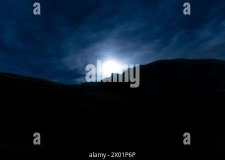 Eine Bergkette hat eine Silhouette vor einem dunklen Himmel mit hellem Mond. Die Szene ist ruhig und friedlich, der Mond strahlt ein sanftes Leuchten auf dem Land aus Stockfoto