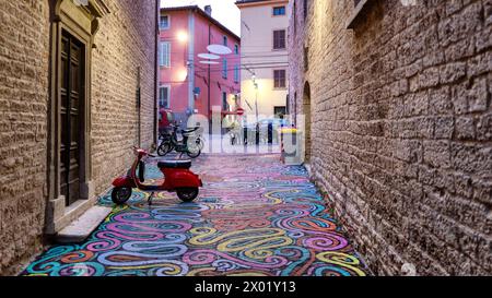 Cagli, Italien - 13. September 2023: Ein roter Roller steht in einer gepflasterten Gasse in Cagli, Italien, wo der Boden mit lebendiger Straßenkunst geschmückt ist. Stockfoto