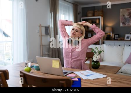 Endbearbeitung. Junge schöne blonde Geschäftsfrau, die zu Hause im Home Office arbeitet, online auf Laptop-Computer arbeitet, sich danach müde und erschöpft fühlt Stockfoto