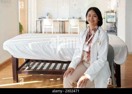 Lächelnder Hausarzt, der vor dem Krankenhausbett auf der Station sitzt Stockfoto