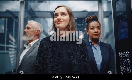 Drei verschiedene internationale Menschen fahren mit einem Glasaufzug zum Büro in einem modernen Business Center. Konzentrieren Sie sich auf eine glückliche junge, schöne Geschäftsfrau, die in einem Aufzug vorne steht. Stockfoto