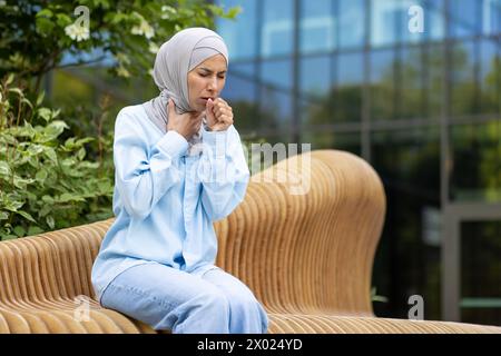 Muslimische Frau hustet und fühlt sich krank, während sie eine Lungenerkrankung hat und draußen sitzt. Kämpfende Dame, die den Mund mit geballter Faust bedeckt, weil sie im Stadtteil keine Viren verbreitet. Stockfoto
