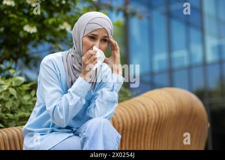 Traurige muslimische Frau, die auf Holzbank sitzt und das Gesicht mit Papiertaschentüchern zum Ausblasen bedeckt. Kranke Frau, die allergisch gegen Pollen ist und versucht, unangenehme Symptome der laufenden Nase zu verstecken. Stockfoto