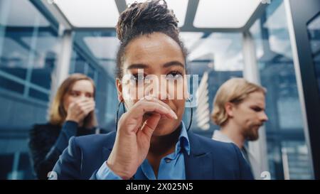 Close Up Porträt einer Frau mittleren Alters schlummert in einem überfüllten Glasaufzug in einem modernen Bürogebäude. Geschäftsfrau bedeckt ihr Gesicht, aber andere Leute haben Angst, den Virus in einem Aufzug zu fangen. Stockfoto