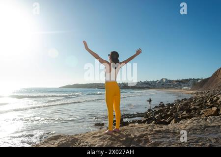 Sportliche Frau, die mit erhobenen Armen am Meer steht und die Sonne genießt, positive Emotionen Stockfoto