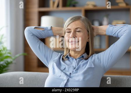 Porträt einer zufriedenen Frau in blauer Bluse, die sich auf der Rückseite des Sofas stützt und mit geschlossenen Augen die Hände hinter dem Kopf hält. Lächelnde Dame, die sich wohlfühlt, während sie sich auf Möbeln in einem stilvollen Apartment entspannt. Stockfoto