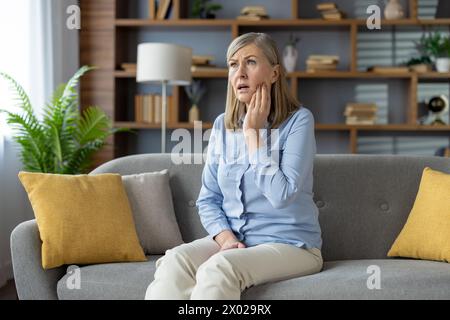 Die reife Dame fühlt Zahnschmerzen und reibt die Wange mit der Hand, während sie mit offenem Mund sitzt. Blonde Frau mit Zahnschmerzen und einem Besuch in der Klinik, um Entzündungen loszuwerden. Stockfoto