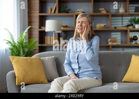 Eine belastete Person, die auf einer grauen Couch sitzt und das Gesicht mit der Hand ergreift, während sie eine Munderkrankung erlebt. Ältere Frau mit Zahnempfindlichkeitskrankheit und unerträglichen Zahnproblemen. Stockfoto