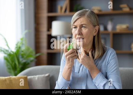 Die kranke Seniorin berührt den Hals und fährt Aerosol in den Mund, während sie sich im Inneren der Wohnung schwer zu atmen fühlt. Asthmatische Frau mit verschreibungspflichtiger Analgetikabehandlung während Allergieanfall. Stockfoto