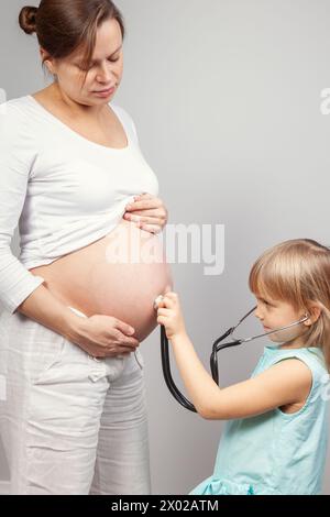 Neugieriges Mädchen, das Stethoskop am Bauch ihrer schwangeren Mutter benutzt, während sie vor neutralem Hintergrund zusammenstehen. Moment der Familienbindung und an Stockfoto