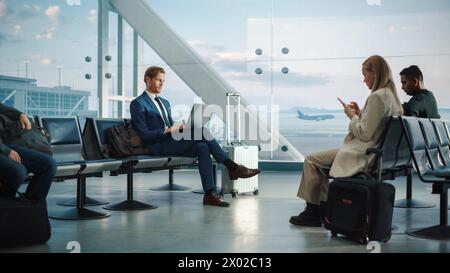 Geschäftiger Flughafenterminal: Gutaussehender Geschäftsmann, der an einem Laptop arbeitet, während er auf seinen Flug wartet. Personen, die in einer Boarding Lounge des Big Airline Hub sitzen und Flugzeuge starten und ankommen Stockfoto