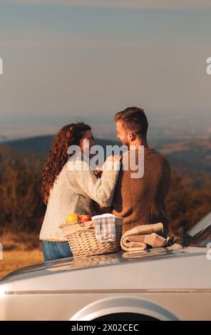 Rückansicht eines Paares, das bei Sonnenuntergang Picknick genießt. Sie sitzen auf einem altmodischen Auto und schauen auf die Distanz. Stockfoto
