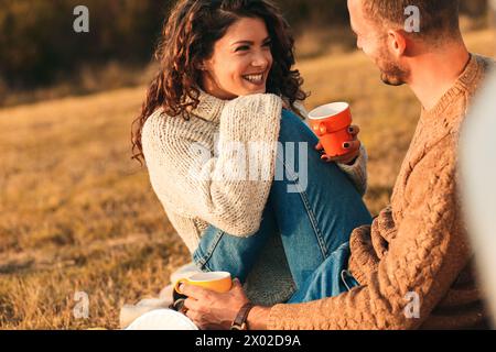 Ein wunderschönes junges Paar, das bei Sonnenuntergang picknickt, Tee trinkt und auf einer Wiese sitzt und sich an ein altmodisches Auto lehnt. Stockfoto