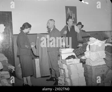 EAST HAM INFORMATION BUREAU: KRIEGSHILFE IN LONDON, 1941 - in einem anderen Teil der East Ham Town Hall kontrollieren Frauen des Women's Voluntary Service (WVS) die kostenlose Ausgabe von Kleidung für die bombardierten Bürger. Hier kann man sehen, wie ein WVS-Mitglied einem Mann mit einer neuen Jacke hilft (es scheint, als ob es einmal eine Army Tunika war), und ein zweites WVS-Mitglied hilft einem anderen Mann mit einer neuen Hose. Im Vordergrund ist ein Stapel von Schuhkartons zu sehen, und eine Kiste mit den Buchstaben RC und einem Kreuz (wahrscheinlich um anzuzeigen, dass der Inhalt vom Roten Kreuz gesammelt wurde) ist ebenfalls zu sehen. Ein Poster an Stockfoto