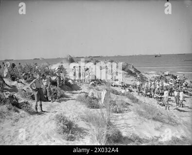 Am 9. Und 10. Juli 1943, während der Operation Husky, waten Truppen in Sizilien an Land und trocknen ihre Kleidung nach der Landung. Die Invasionsflotte ist im Hintergrund sichtbar. Stockfoto