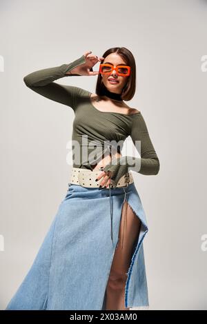 Eine modische junge Frau mit brünetten Haaren, die selbstbewusst in einem Rock und einer Sonnenbrille in einem Studio-Setting posiert. Stockfoto