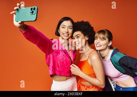 Drei Frauen, die verschiedene Kulturen repräsentieren, genießen einen spielerischen Moment, während sie ein Selfie mit einem Handy vor orangem Hintergrund machen. Stockfoto