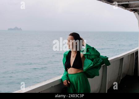 Lächelnde Frau steht auf einem Fährdeck, ihre Haare werden vom Meereswind zerzaust, mit bewölktem Himmel und fernem Land Stockfoto